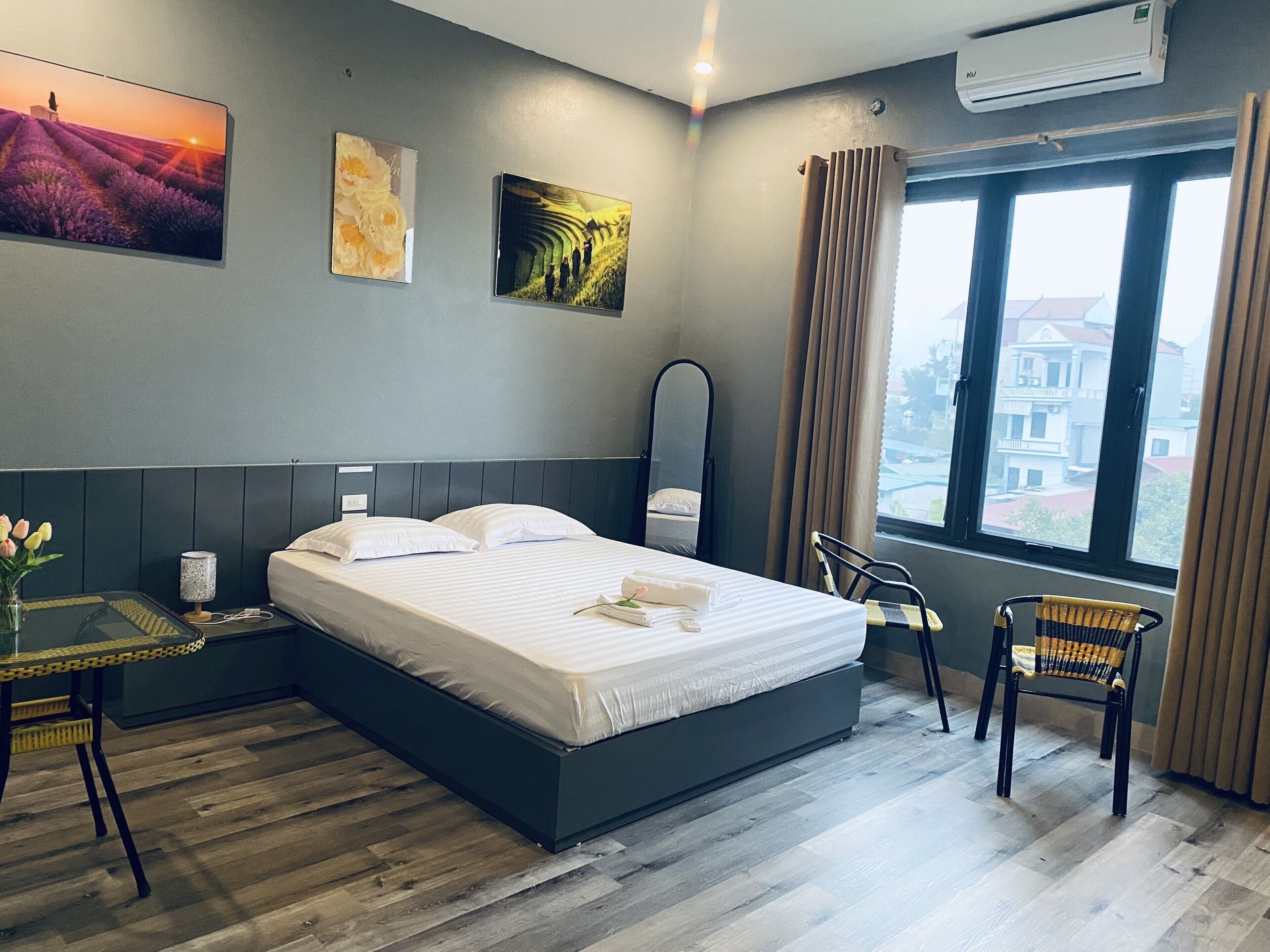Une chambre d’hôtel avec un grand lit, une table de chevet avec une lampe, une chaise, une fenêtre avec des rideaux et des œuvres d’art accrochées au mur.