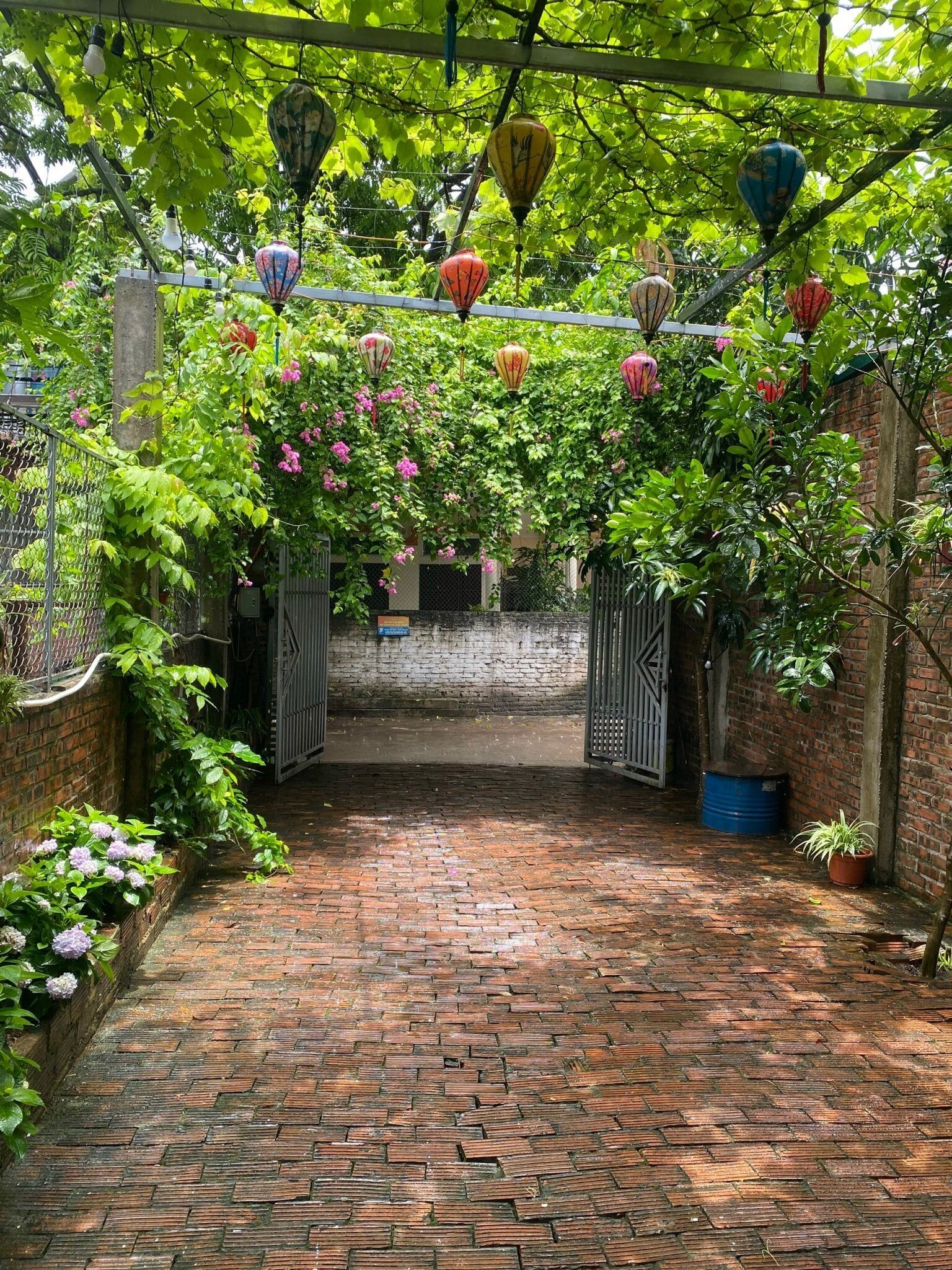 Een met stenen bestraat pad dat door een tuin leidt, met een metalen hek, omringd door weelderig groen en lantaarns.