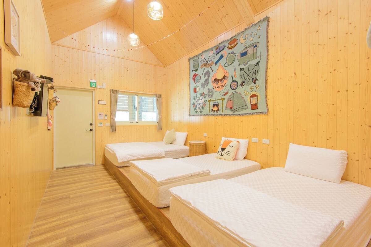 Une chambre de style cabane avec des murs et des planchers en bois, trois lits simples, une grande tapisserie sur le thème du camping et une petite fenêtre avec des rideaux.