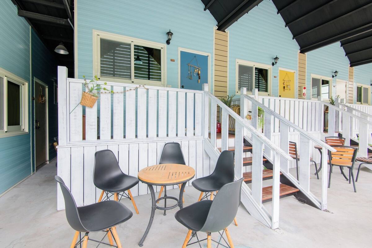 Un espace extérieur aménagé, abrité, avec une table en bois et des chaises noires, une clôture blanche en bois et un escalier menant à un niveau supérieur.