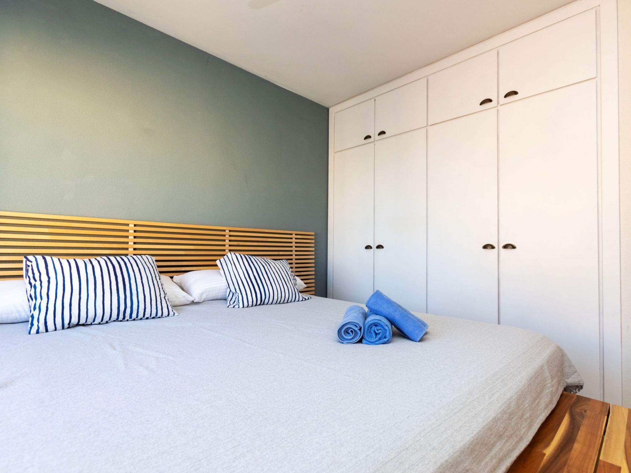 Una cama bien hecha con almohadones a rayas, un cabecero de madera y un armario blanco con varias puertas.