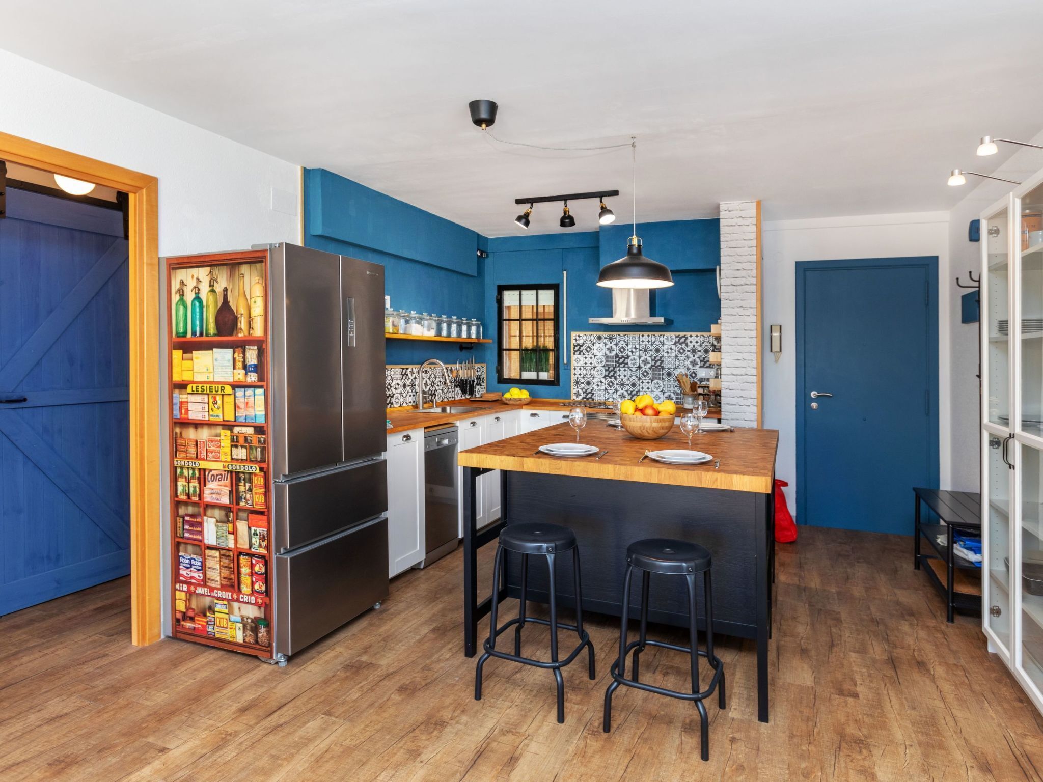 Une cuisine moderne avec des murs bleus, un îlot central en bois, des tabourets noirs et un réfrigérateur en acier inoxydable.
