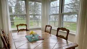 Una mesa de comedor de madera con un centro de mesa floral, rodeada de sillas, frente a una ventana con vistas a árboles y césped.