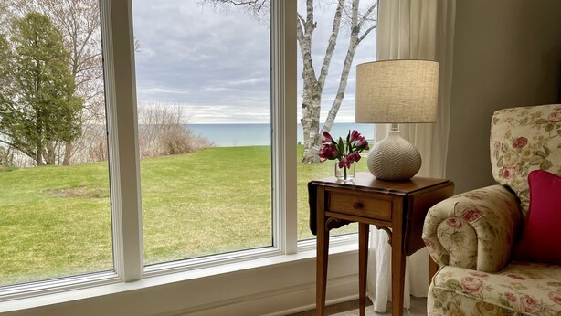 Una habitación con un ventanal grande, un sillón floral, una mesita de noche con una lámpara y un jarrón con flores.
