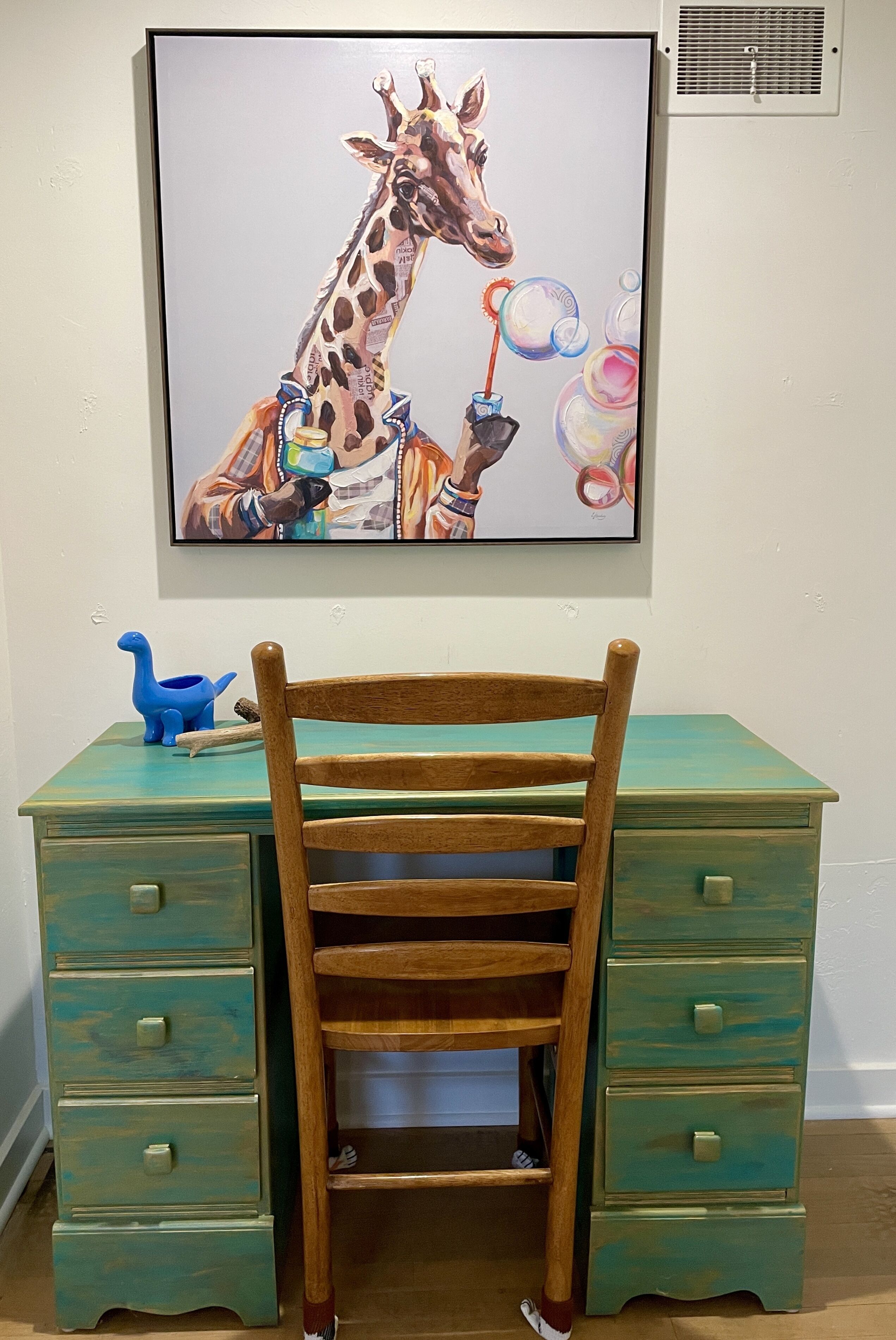A teal-painted desk with a wooden chair, a framed painting of a giraffe, and a blue dinosaur toy.
