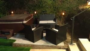An outdoor seating area with a hot tub, chairs, and tables, surrounded by greenery and illuminated by wall-mounted lights. - Family Fun Home (Beverly Hills)