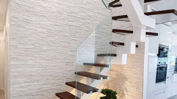 Una escalera moderna con barandillas de vidrio y peldaños de madera, que conduce a una zona de cocina.