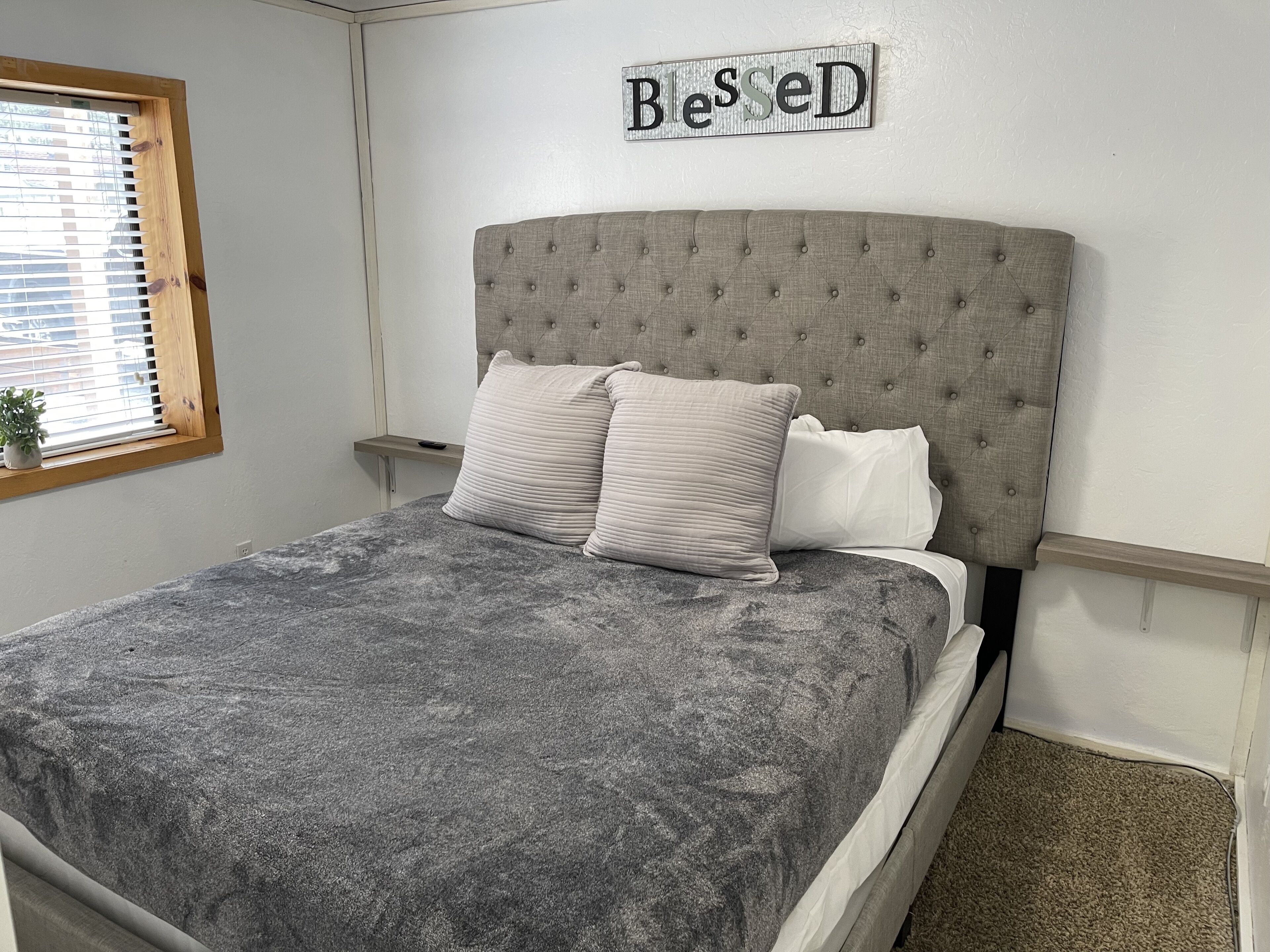 Una cama bien hecha con una colcha gris con textura y dos almohadas, un cartel en la pared que dice "Blessed" y una ventana con persianas.
