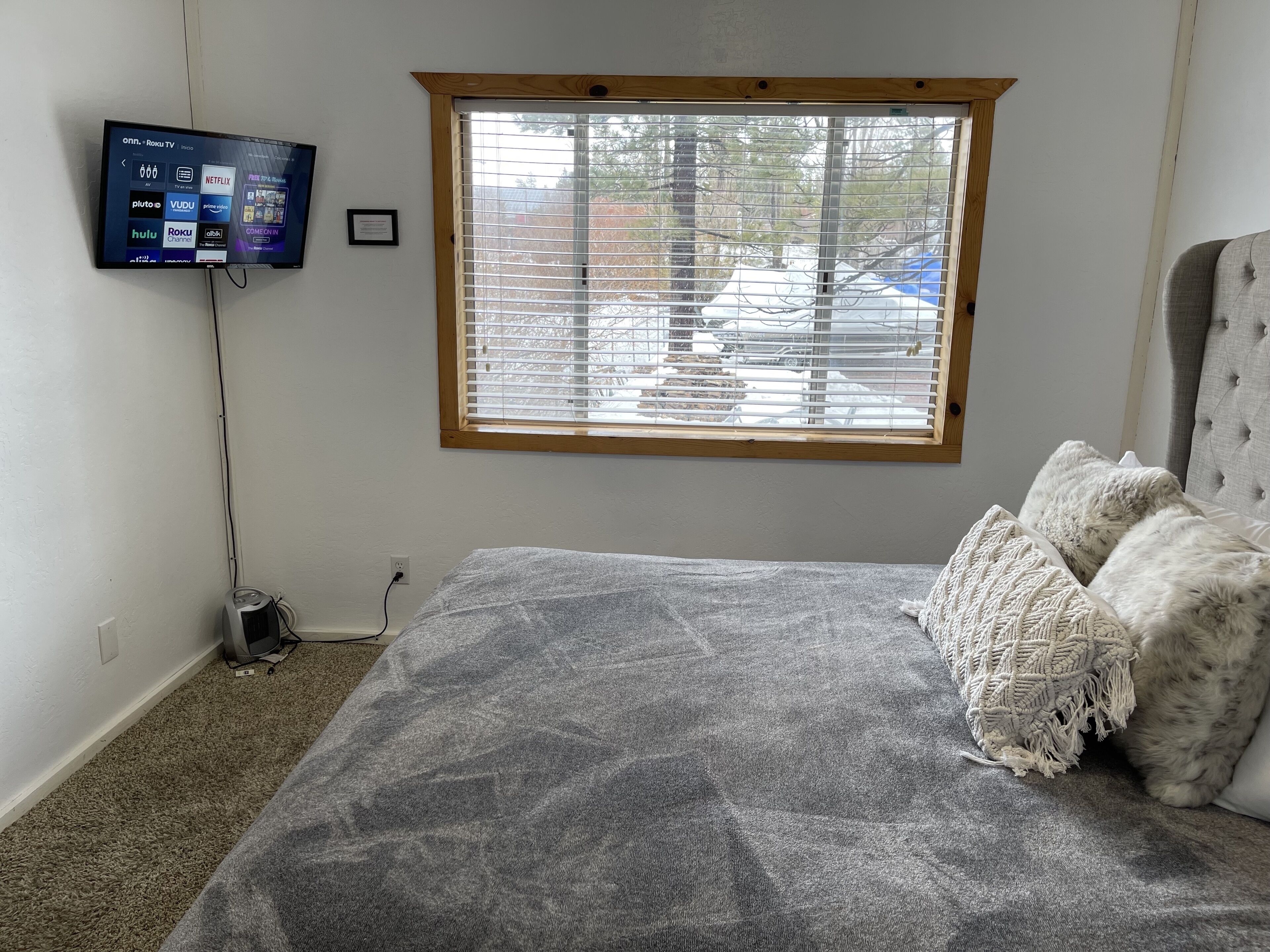 Un dormitorio con cama, televisión montada en la pared, ventana con persianas y piso alfombrado.