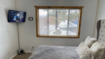 Un dormitorio con cama, televisión montada en la pared, ventana con persianas y piso alfombrado.