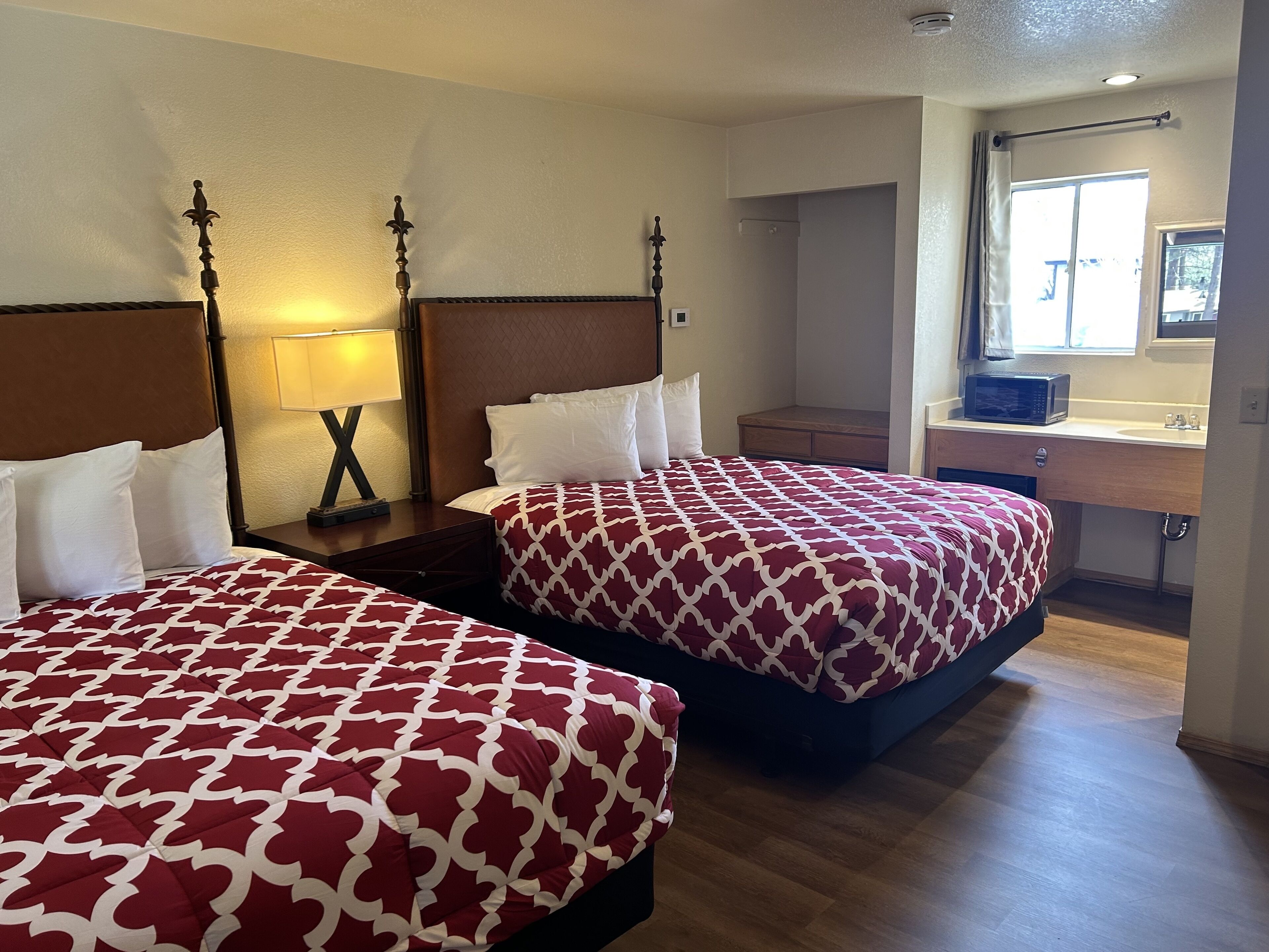 A hotel room with two beds, a nightstand with a lamp, a wooden headboard, a small bathroom with a sink and mirror, and a window with curtains.
