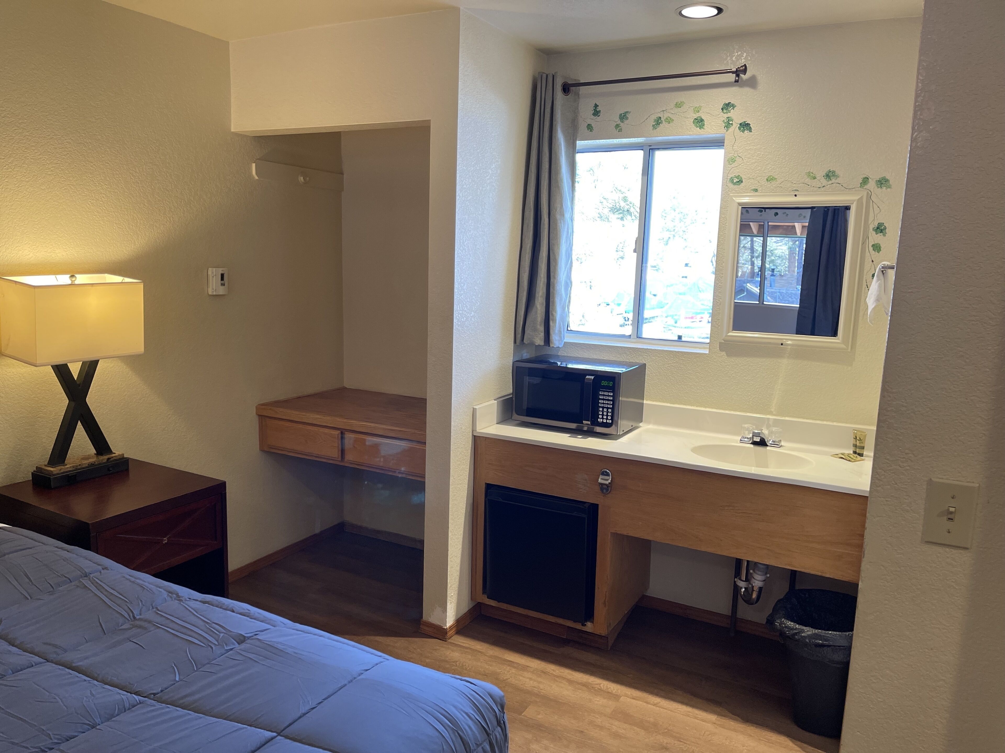 Habitación de hotel pequeña con cama, escritorio con microondas, lavamanos y cubo de basura.