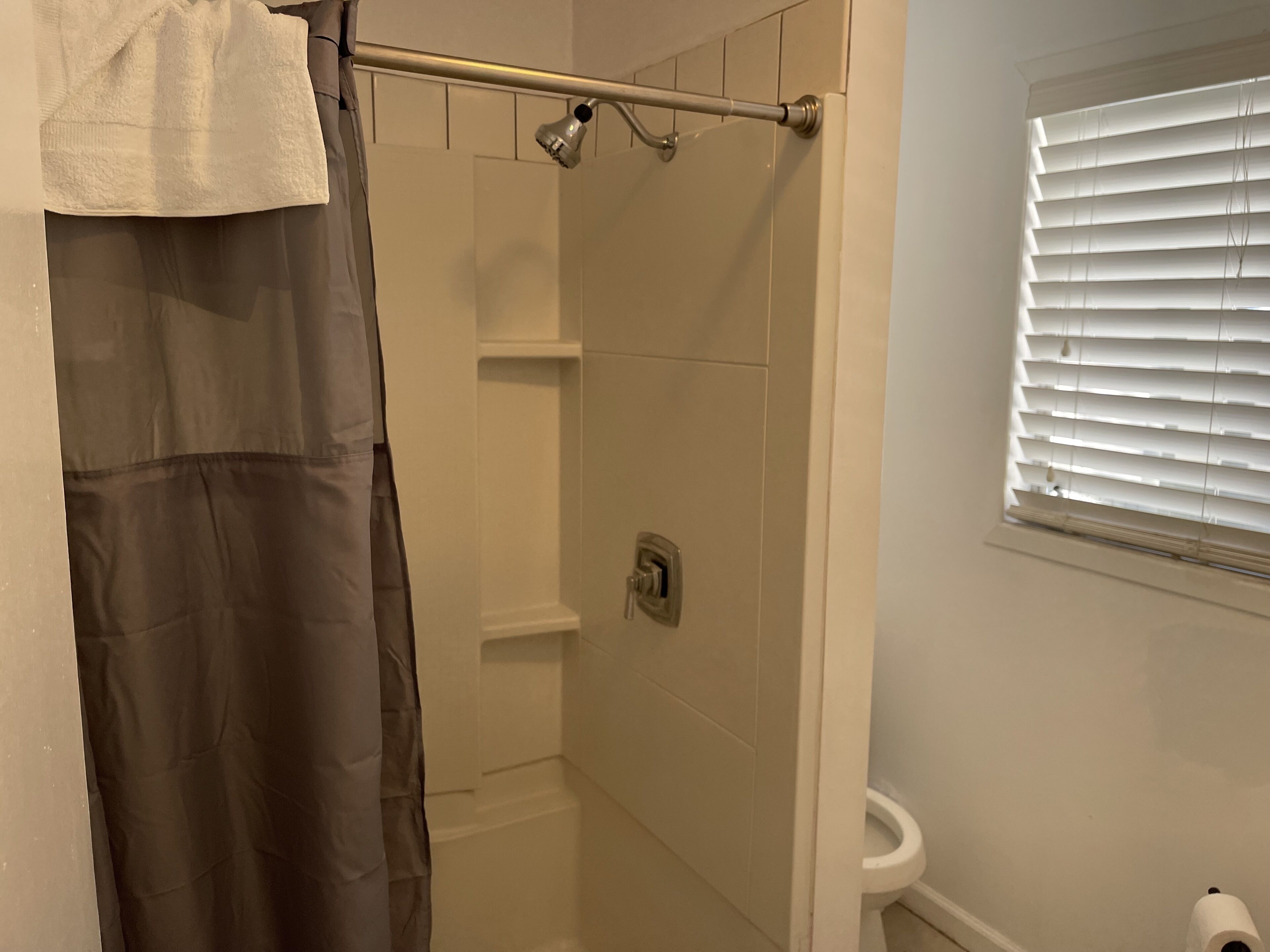 A bathroom with a shower, toilet, and window with blinds.