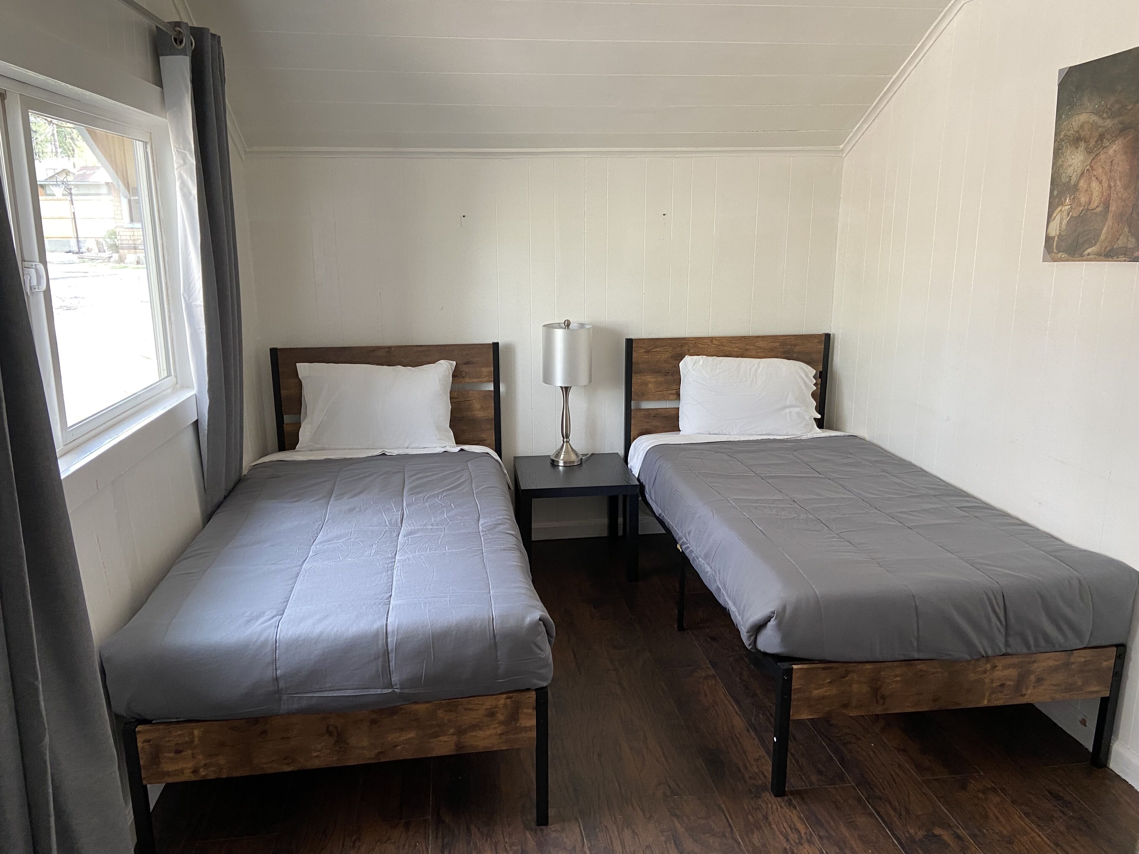 A room with two single beds, a nightstand with a lamp, wooden flooring, and a window with curtains.