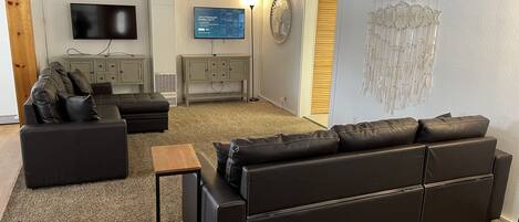 A living room with a large black leather sofa, a smaller black sofa, a wooden coffee table, a flat-screen TV mounted on the wall, and a decorative wall hanging.