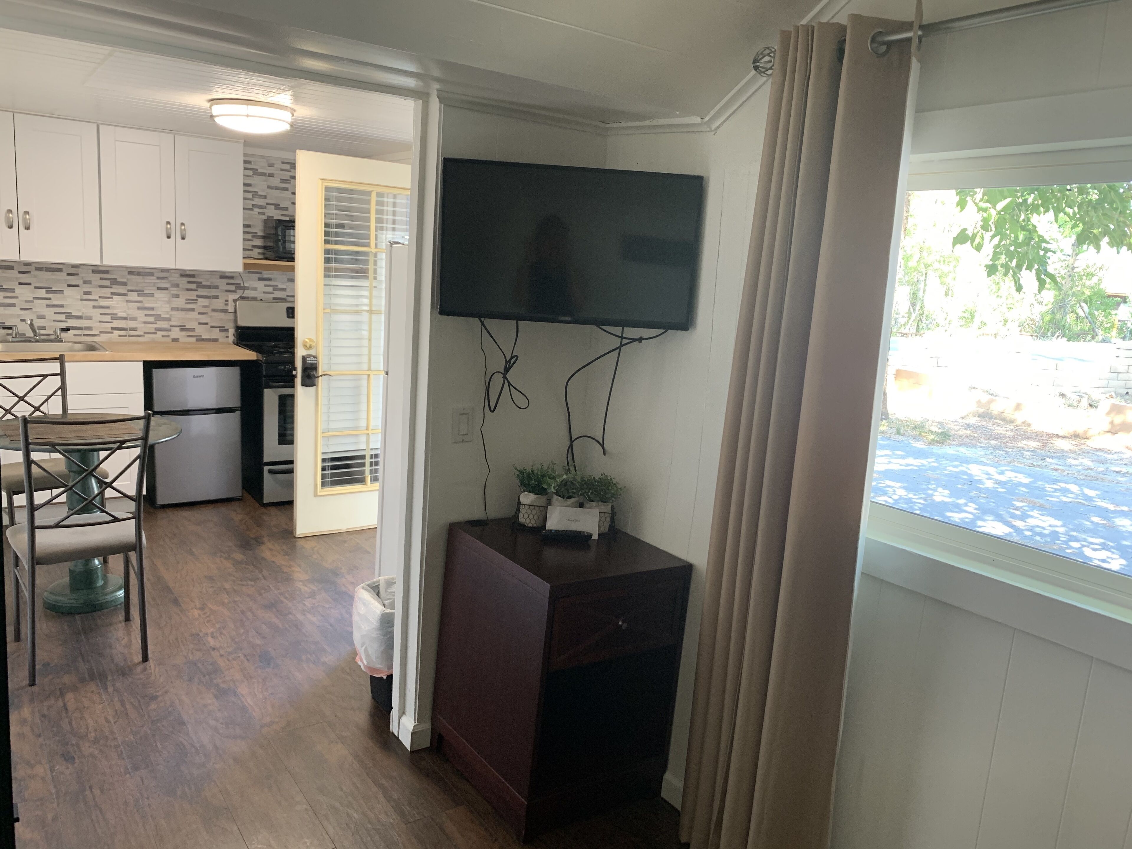 A compact kitchen with white cabinets, a built-in oven, and a refrigerator. A flat-screen TV is mounted on the wall, and there's a small table with chairs.