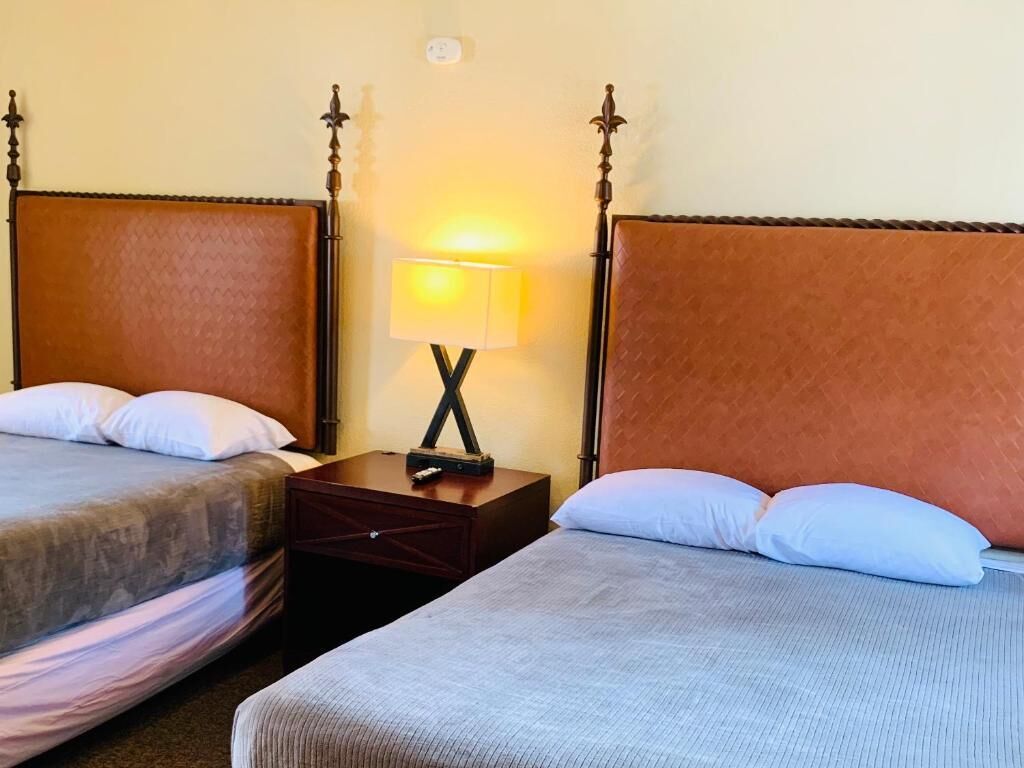 A hotel room with two beds, a nightstand with a lamp, and a wall-mounted light fixture.