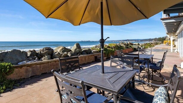 An outdoor seating area with tables and chairs, a large umbrella, and a view of the ocean.