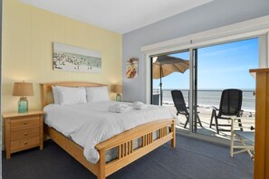 A hotel room with a large bed, a nightstand, a lamp, a painting, and a view of the beach through a sliding glass door. - Beachfront Rio Del Mar Home - 5 bedrooms! (Aptos)