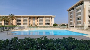 A swimming pool surrounded by lush greenery, with a multi-story building featuring balconies in the background. - Port-Fréjus, Cesaree Borghèse, 2/3 rooms 52m2, 4 beds, swimming pool, air-conditioned, parking, close access to the beach, large balcony for a pleasant stay in the sun, relaxation and leisure (Fréjus)