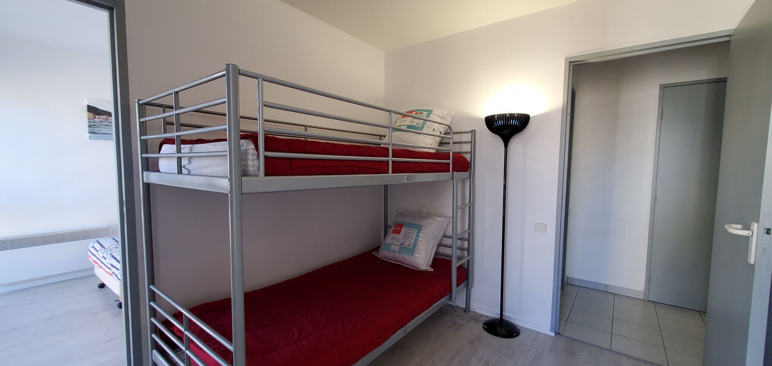 Habitación con literas, un colchón rojo, una lámpara de pie y una puerta.