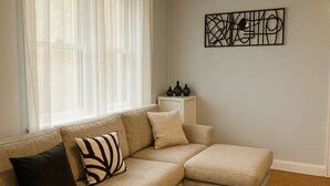 A living room with a beige sectional sofa, a patterned rug, and a wall art piece. - Family-Friendly 4BR 10min MetLife & American Dream Mall• 20min NYC •Free Parking (North Bergen)