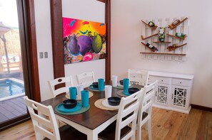 Un comedor con una mesa de madera, sillas blancas y un cuadro colorido en la pared.