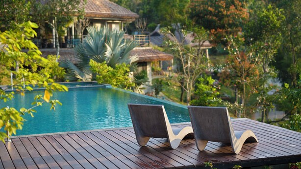 A wooden deck with two lounge chairs facing a swimming pool, surrounded by lush greenery and a house with a thatched roof. - Blanket Days Resort and Spa (Peermade)