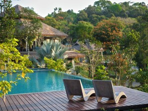 Una terraza de madera con dos sillas reclinables orientadas a una piscina, rodeada de exuberante vegetación y una casa con techo de paja.