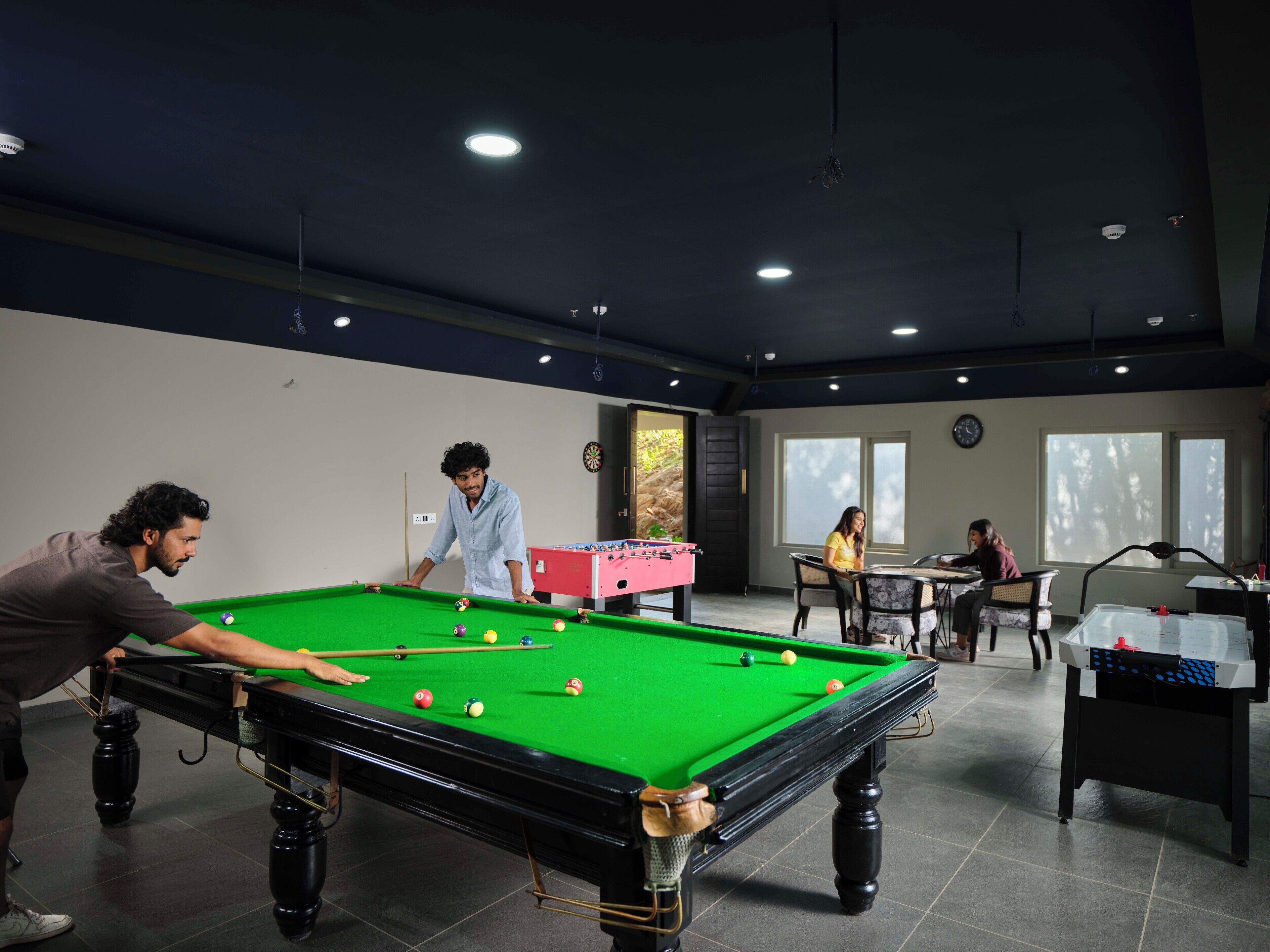 Une table de billard avec des joueurs, une table de baby-foot et un jeu de fléchettes dans une pièce dotée de grandes fenêtres et d’une horloge au mur.