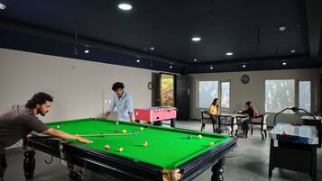 Une table de billard avec des joueurs, une table de baby-foot et un jeu de fléchettes dans une pièce dotée de grandes fenêtres et d’une horloge au mur.