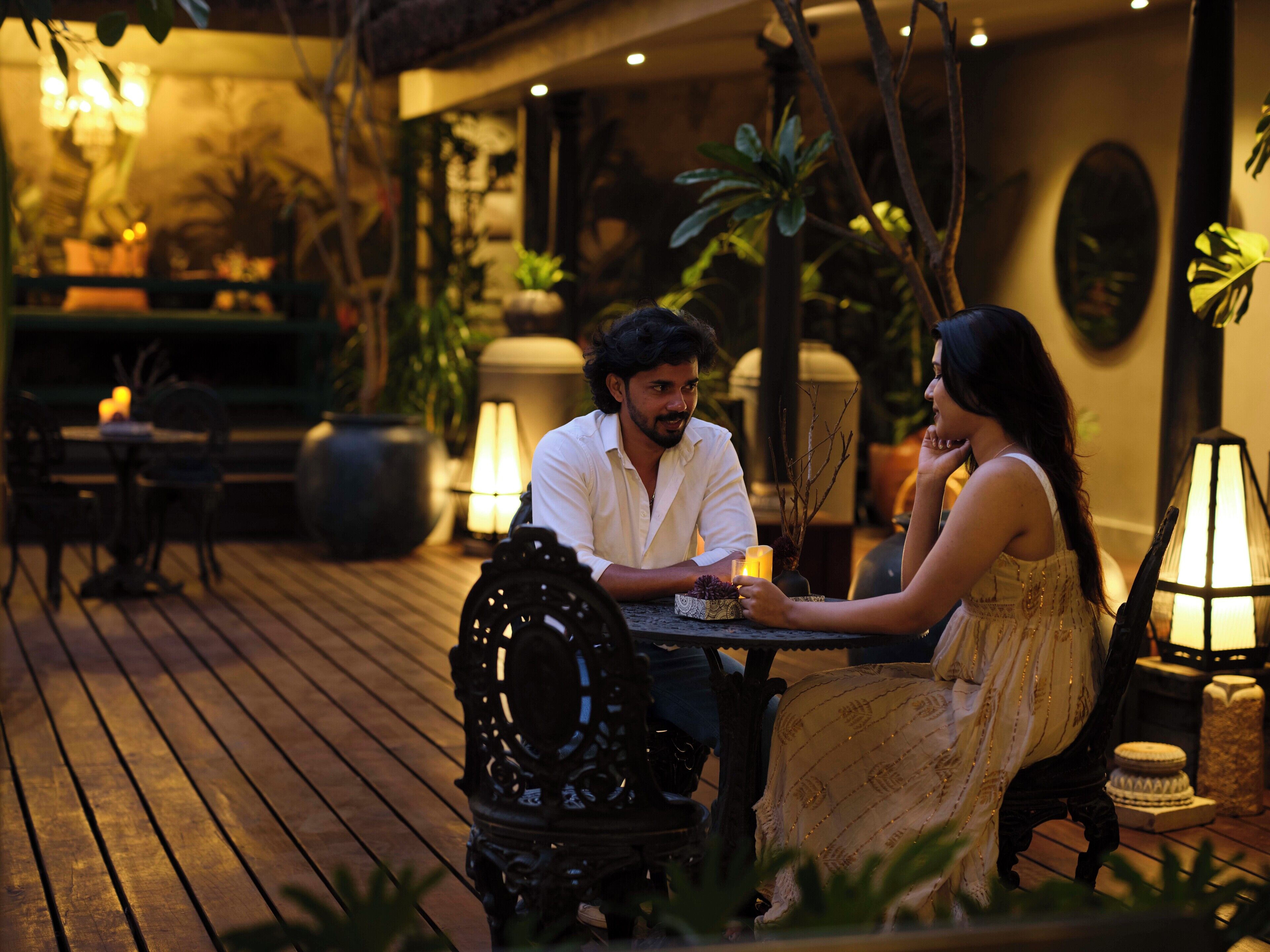 Un couple est assis à une table à l’extérieur, éclairée par des bougies, et est entouré de plantes en pot et d’une terrasse en bois.