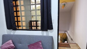 A room with a grey sofa, two purple pillows, a window with black curtains, and a wooden floor.