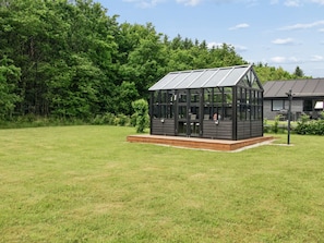 A glass conservatory with a wooden deck, surrounded by a grassy field and trees.