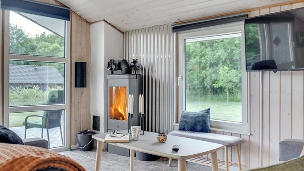 A cozy room with a fireplace, a table with a remote control, and a large window offering a view of greenery.