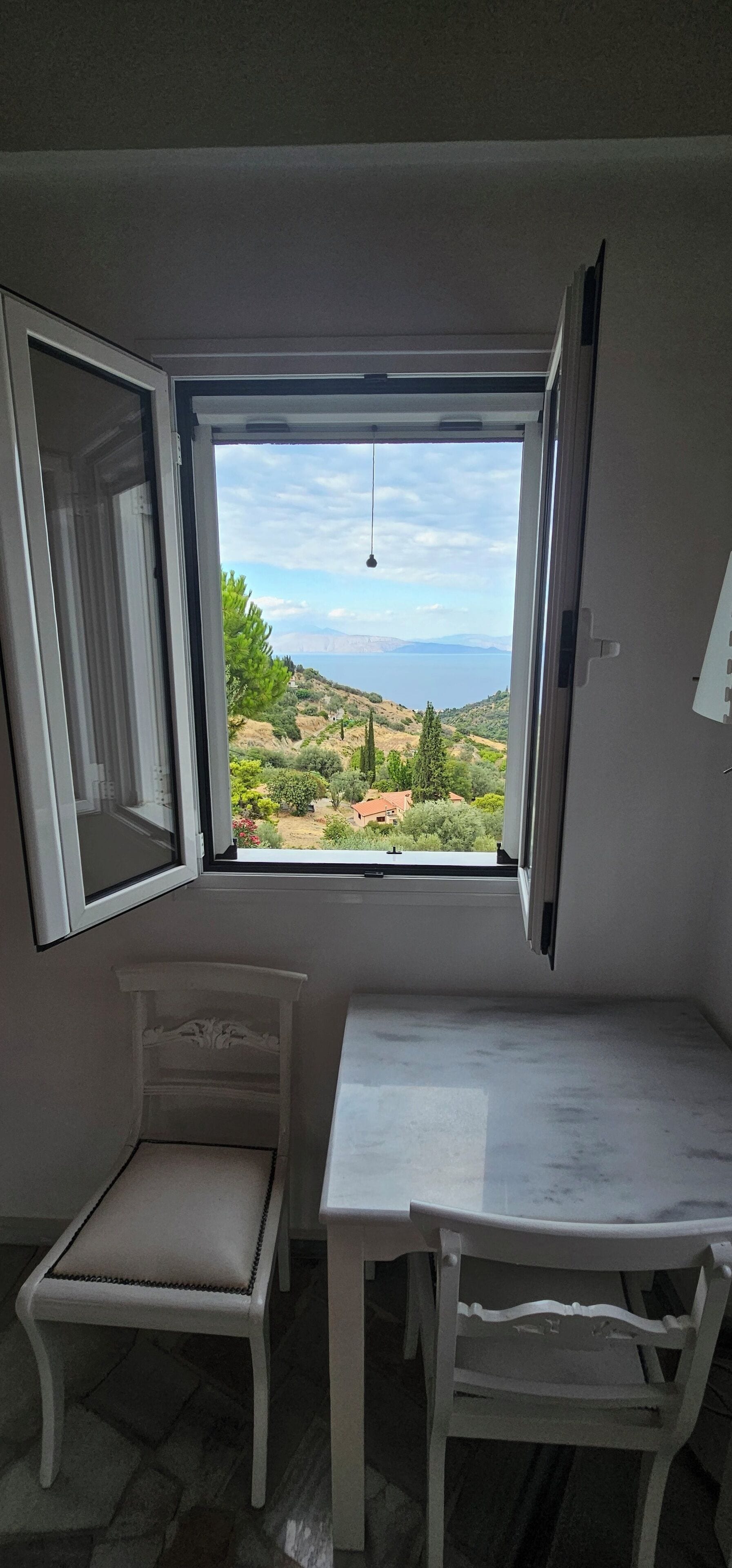 A room with a table and a chair, a window with an open shutter, and a view of a landscape with trees and a body of water.