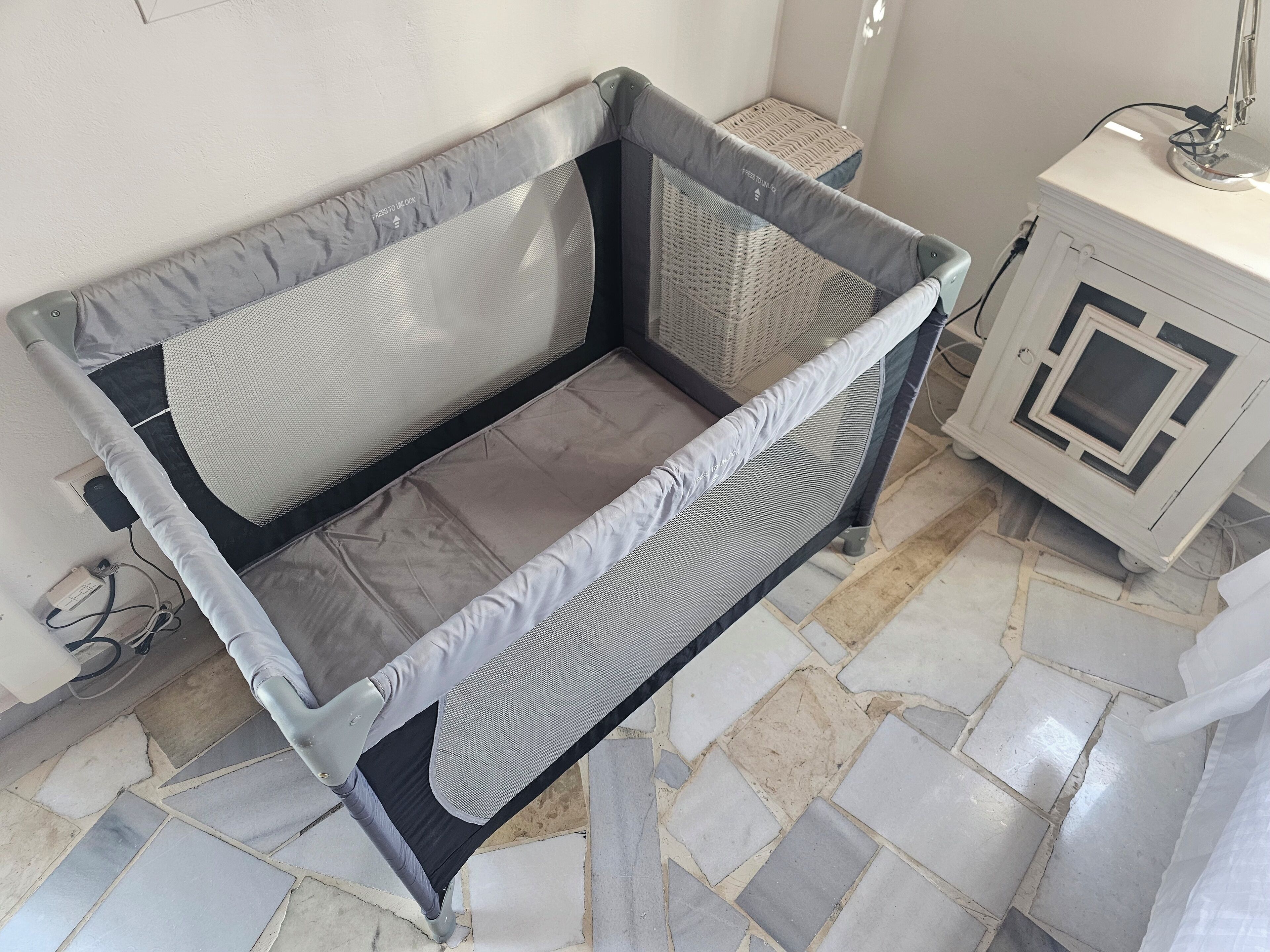 A room with a bed covered in a grey mattress protector, a small bedside table with a lamp, and a tiled floor.