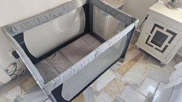 A room with a bed covered in a grey mattress protector, a small bedside table with a lamp, and a tiled floor.