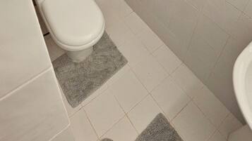 Un bagno piccolo e funzionale con WC, bidet e lavabo. Le pareti sono rivestite di piastrelle e sopra il WC c’è un pannello in legno.