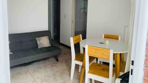 En kompakt stue med åbent planløsning, med sofa, spisebord med stole, og et køkken der kan anes gennem en åben dør.