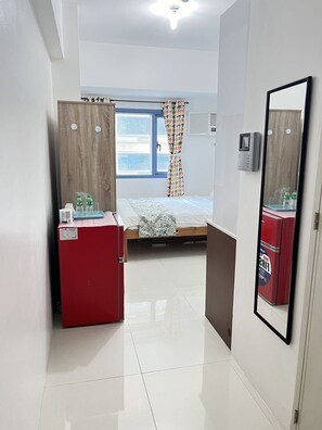 A compact hotel room with a bed, a small desk with a refrigerator, a window with curtains, and a mirror on the wall.