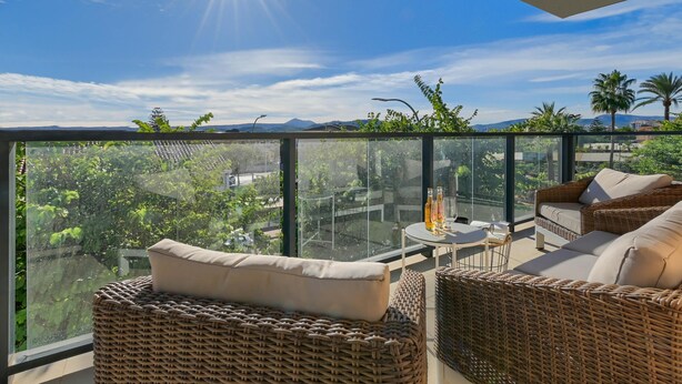 A balcony with wicker furniture, a glass railing, and a view of greenery and distant mountains.