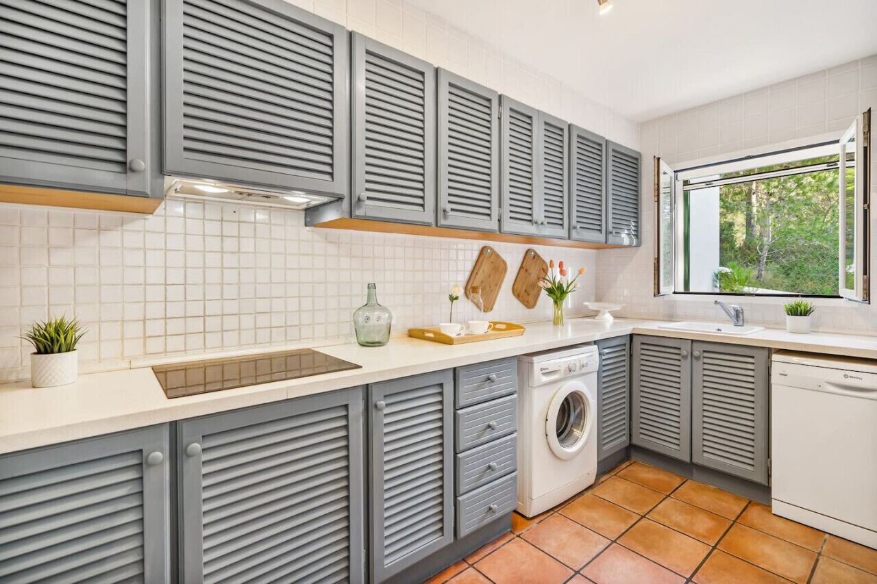 A modern kitchen with grey cabinetry, a built-in washing machine, and a window providing a view of greenery.