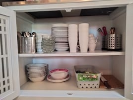 Un mueble de cocina con estantes blancos que contienen diverso menaje, incluyendo platos, tazas y utensilios.