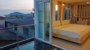 A bedroom with a bed, a glass door leading to a balcony, and a view of buildings and hills. - Habor House by Koh Larn Riviera (Koh Lan)