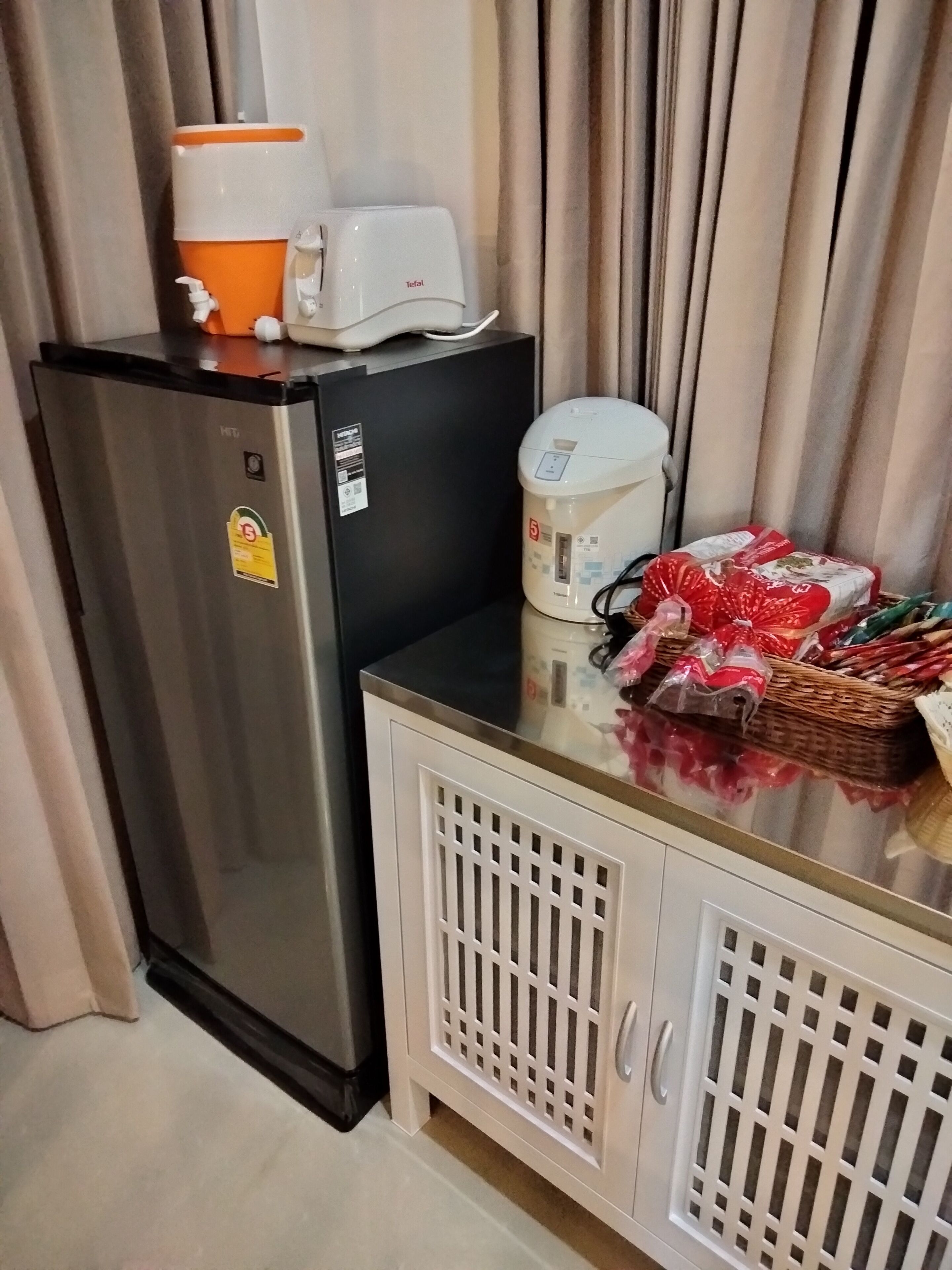 A compact kitchen with a refrigerator, a coffee maker, a kettle, and a basket of groceries on the counter.