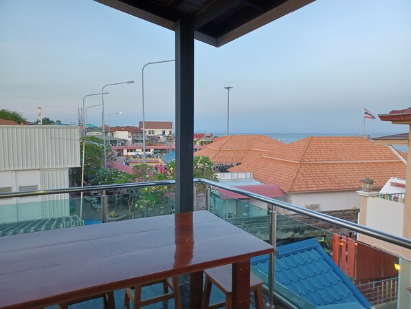 A balcony with a wooden table and chairs, overlooking a town with buildings and a view of the sea.