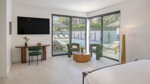 Una habitación de hotel moderna con un televisor grande, una mesa de comedor con sillas, una mesa redonda de centro, una cama y vistas a una piscina y sillas de descanso afuera.