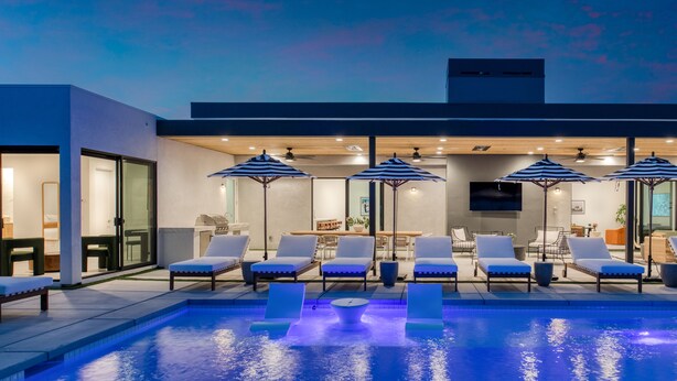 Un área de piscina moderna al aire libre con sillones de descanso y sombrillas, un espacio de estar techado y una vista despejada de la piscina.