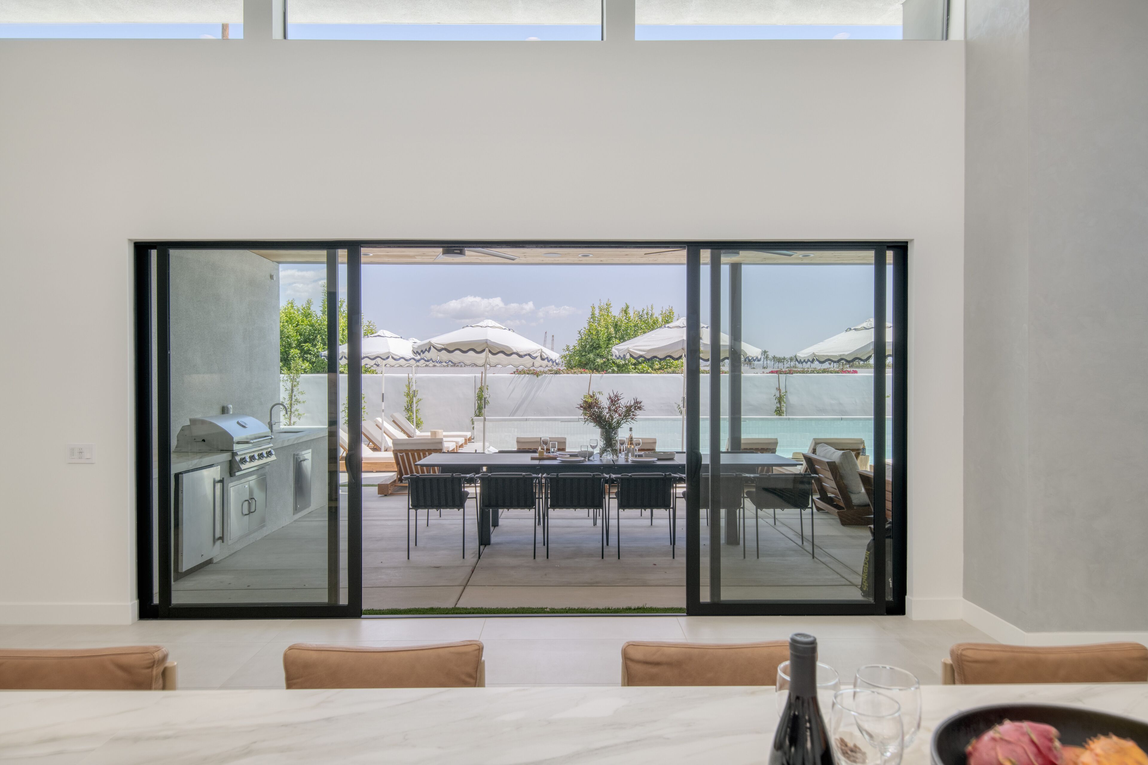 A modern kitchen with a built-in grill, a dining area with a table and chairs, and a large sliding glass door leading to an outdoor patio with umbrellas and a pool.